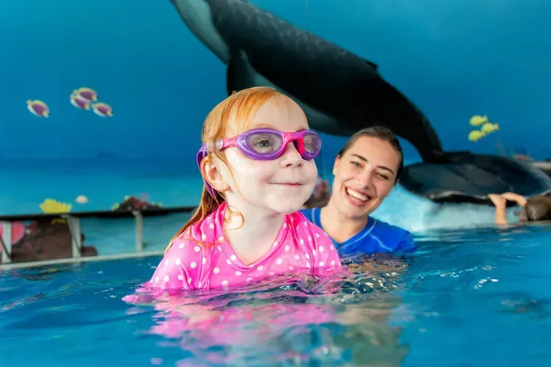 Girl in Pink Swim Shirt and Goggles in Pool with Instructor Under Whale Mural — Swim Care Boambee in Coffs Harbour, NSW