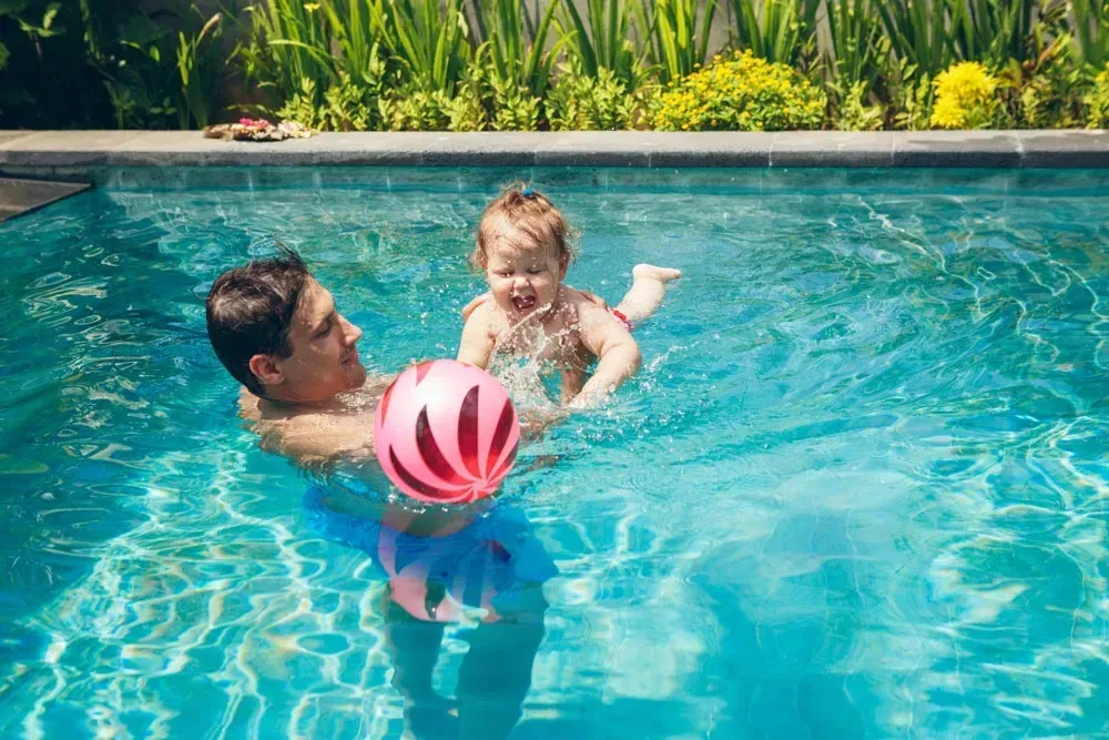 Man and Baby Splashing in Pool with Pink and Blue Ball — Swim Care Boambee in Coffs Harbour, NSW