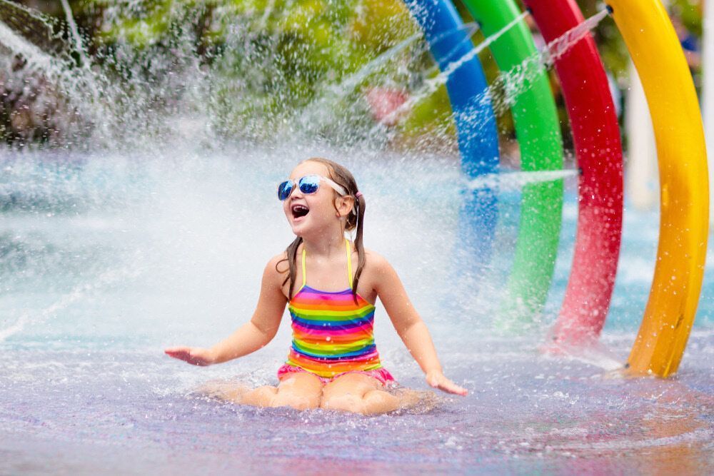 Girl in Sunglasses and Rainbow Swimsuit Plays in Water Fountains — Swim Care Boambee in Coffs Harbour, NSW