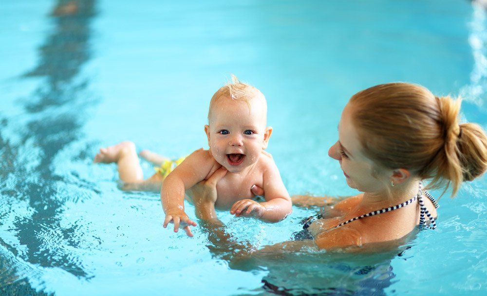 Woman Holding a Baby in A Swimming Pool. the Baby Smiles, Looking at The Camera — Swim Care Boambee in Coffs Harbour, NSW