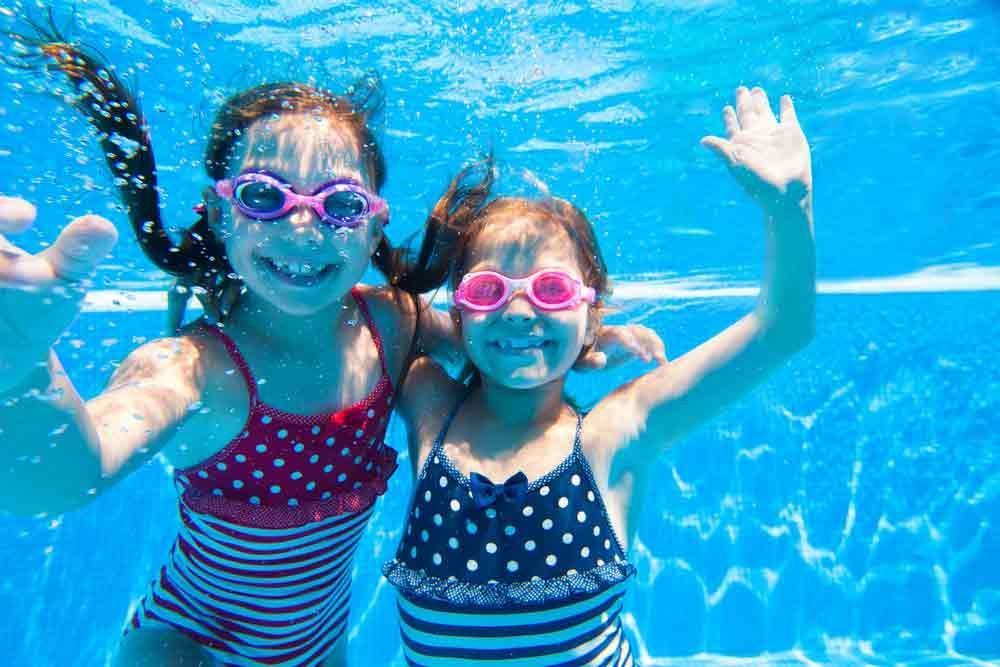 Two Girls in Swimsuits and Goggles Underwater, Smiling and Waving in A Blue Pool — Swim Care Boambee in Coffs Harbour, NSW
