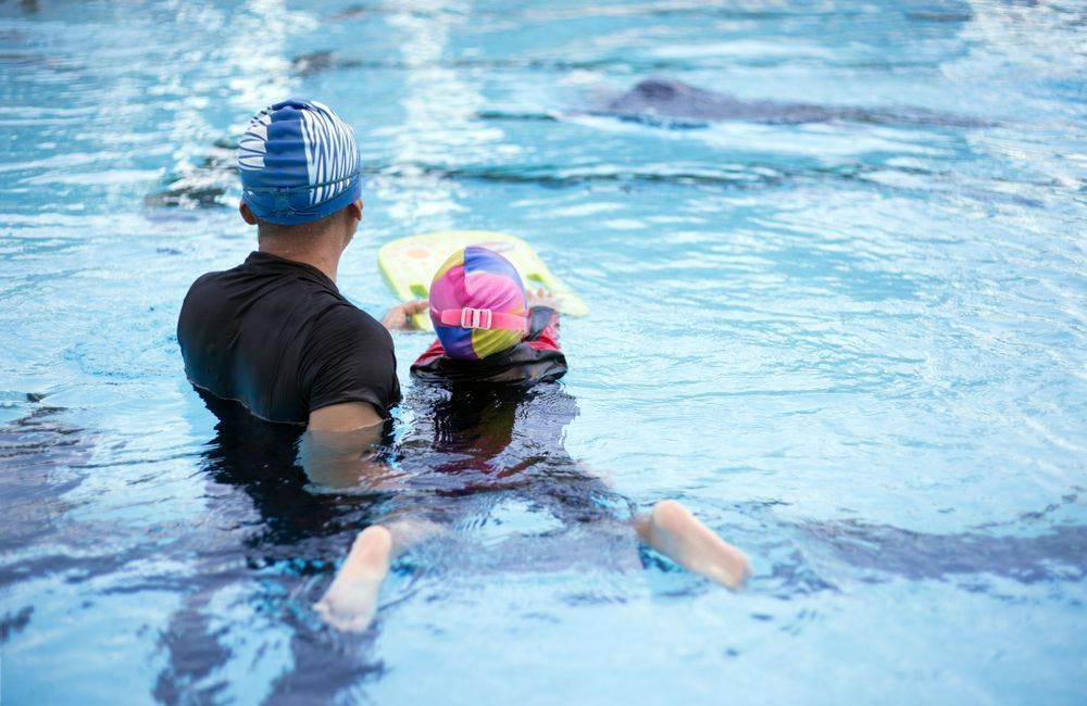 Adult Assisting a Child in A Swimming Pool; Both Are Wearing Swim Caps, Child Holding a Float, Learning to Swim — Swim Care Boambee in Coffs Harbour, NSW