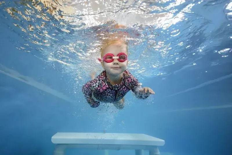 Girl with Red Goggles Swims Underwater in A Blue Pool, Smiling — Swim Care Boambee in Coffs Harbour, NSW