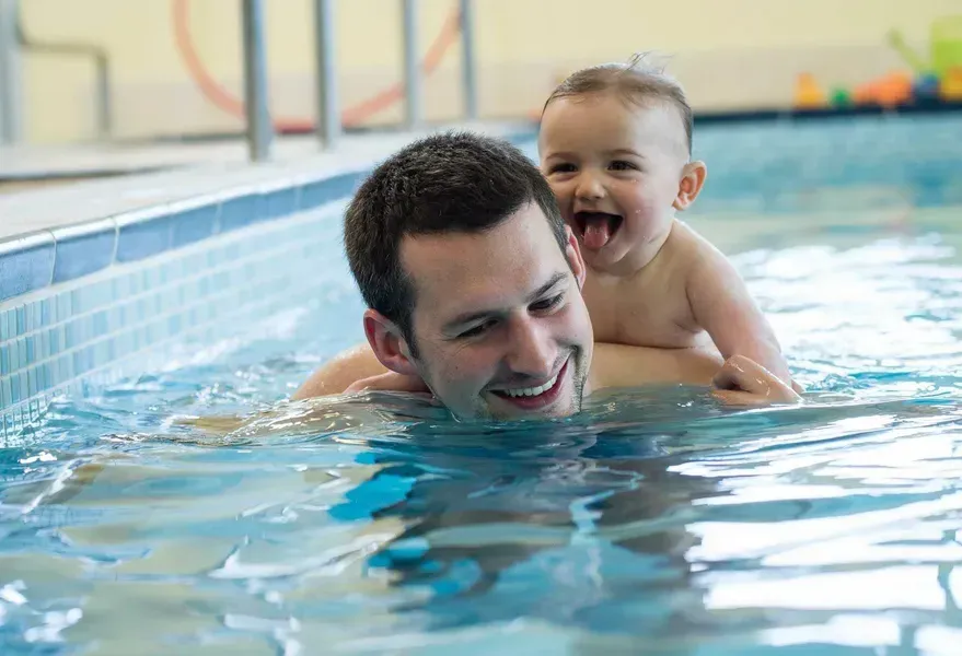 Man in Pool with Smiling Baby on His Back. Blue Pool, Natural Light — Swim Care Boambee in Coffs Harbour, NSW