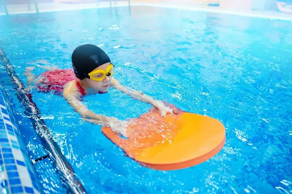 Child in a Pool With a Swim Board, Wearing Goggles and a Cap, Learning to Swim — Swim Care Boambee in Boambee, NSW