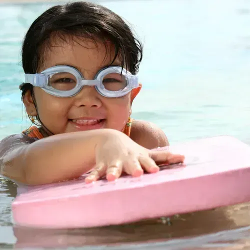 Girl in Goggles Smiles in A Pool, Holding a Pink Kickboard — Swim Care Boambee in Coffs Harbour, NSW