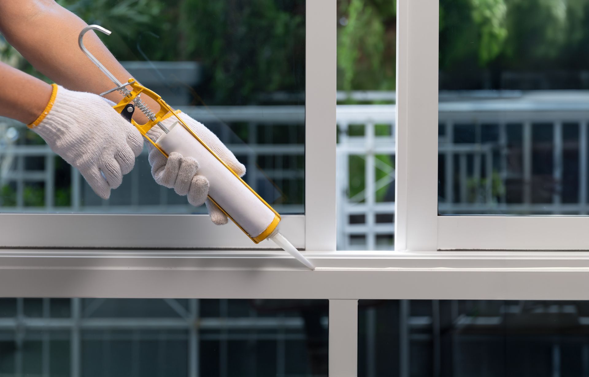 A worker with gloved hands applying silicone sealant caulk to repair a leak inside a window frame.
