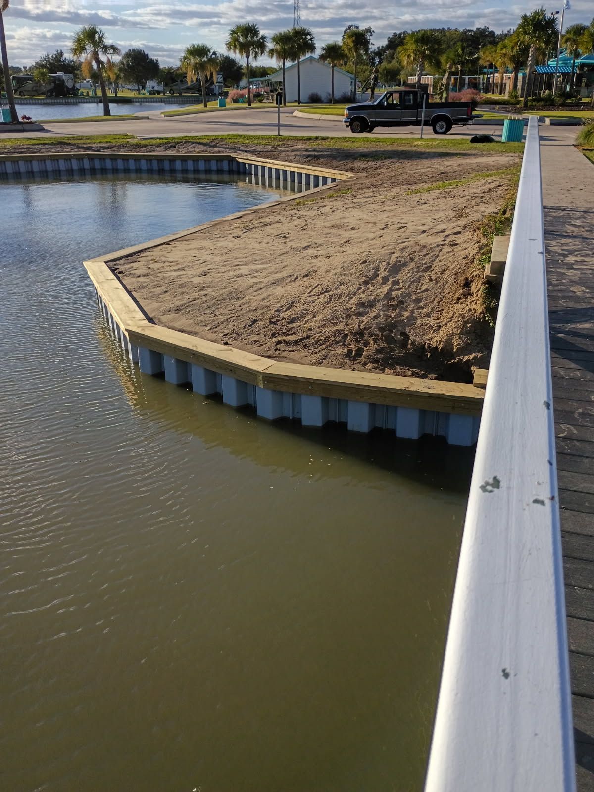A view of a waterfront bulkhead made of white, corrugated panels bordering a sandy land area beside a murky pond.