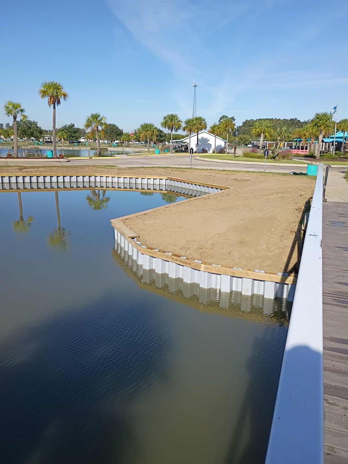 A newly installed grey vinyl bulkhead forms a rectangular corner along a calm lake, with brown sand fill in the foreground.