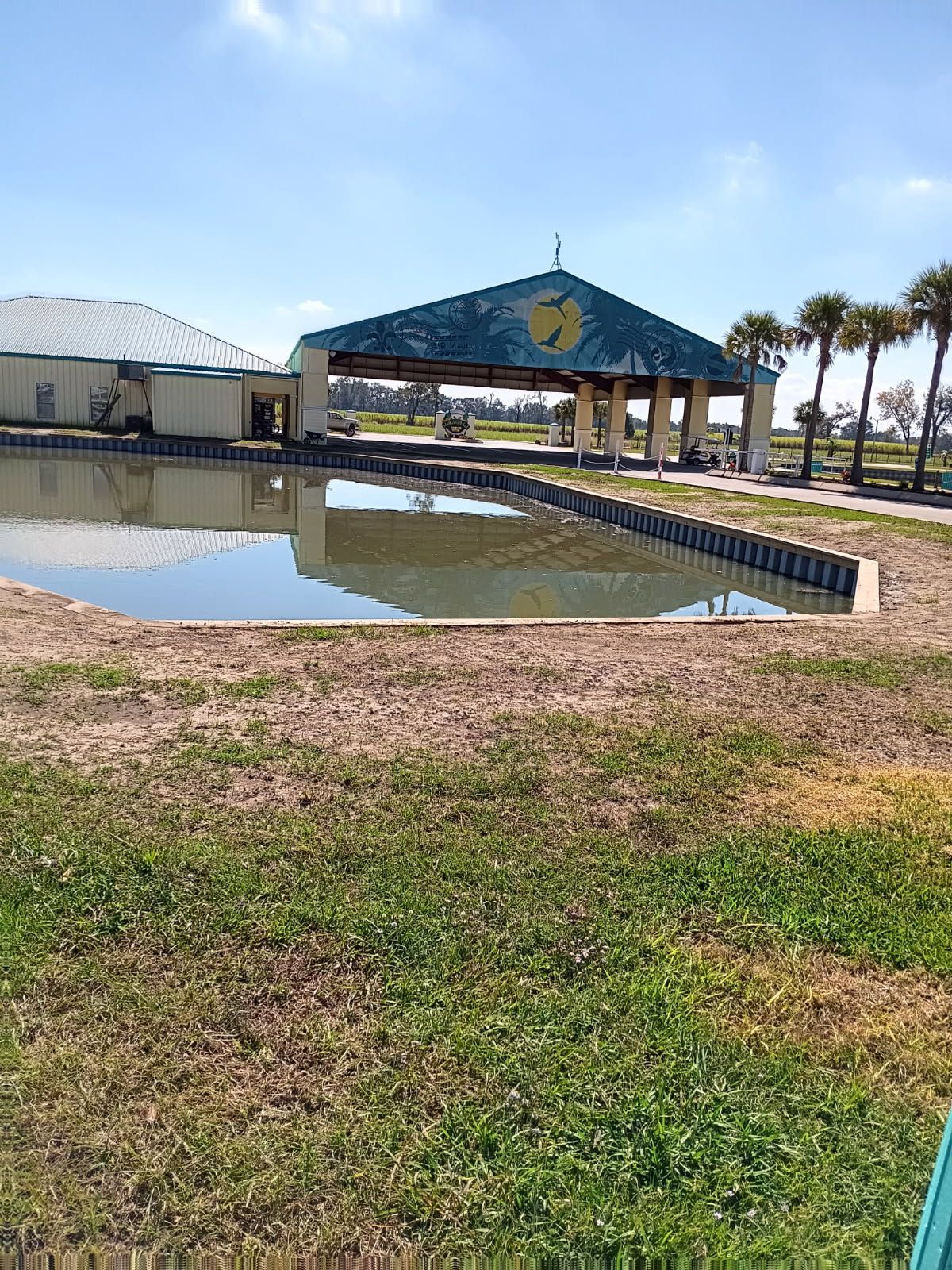 A pond sits in the foreground before a pavilion structure with a teal-colored, peaked roof under a blue sky.