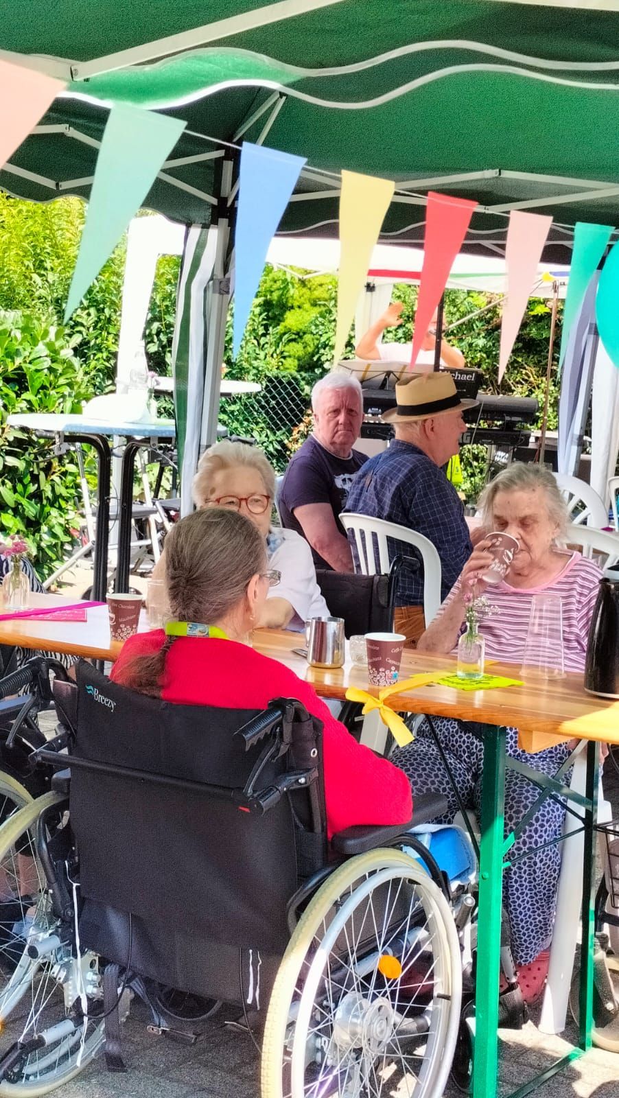 Eine Gruppe von Menschen im Rollstuhl sitzt an Tischen unter einem Sonnenschirm.