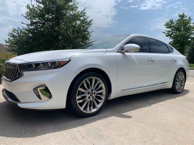 A white car is parked in a driveway with trees in the background.