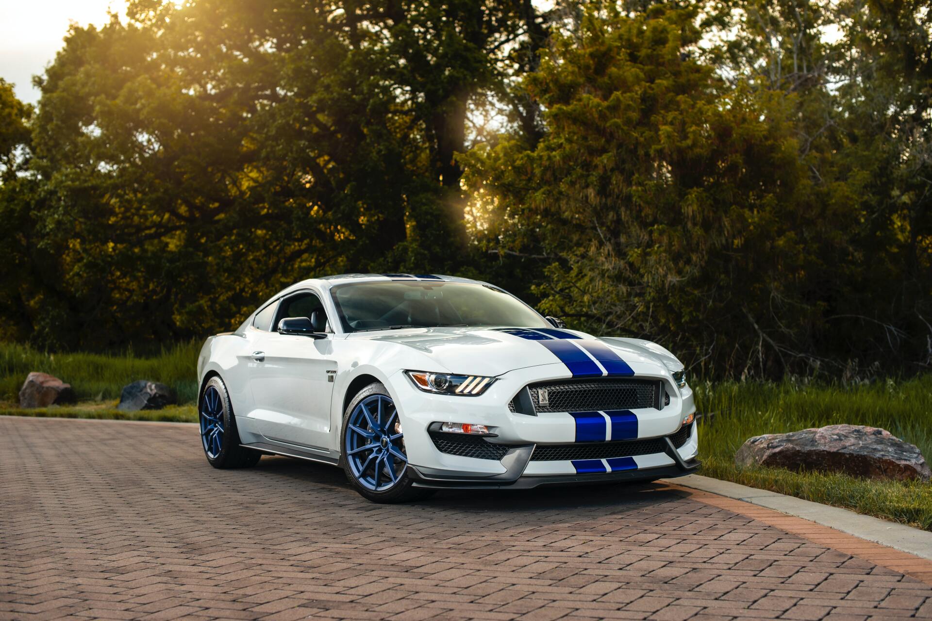 A white ford mustang with blue stripes is parked on a brick road.