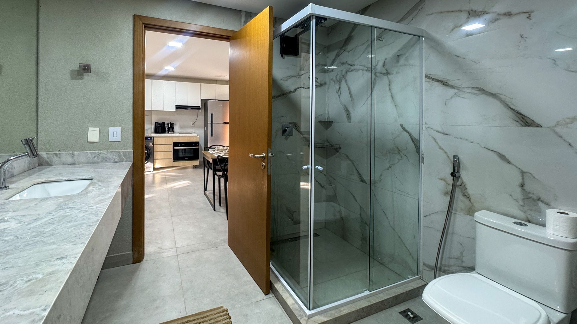 Banheiro com paredes de mármore, chuveiro, vaso sanitário e porta aberta para a cozinha.