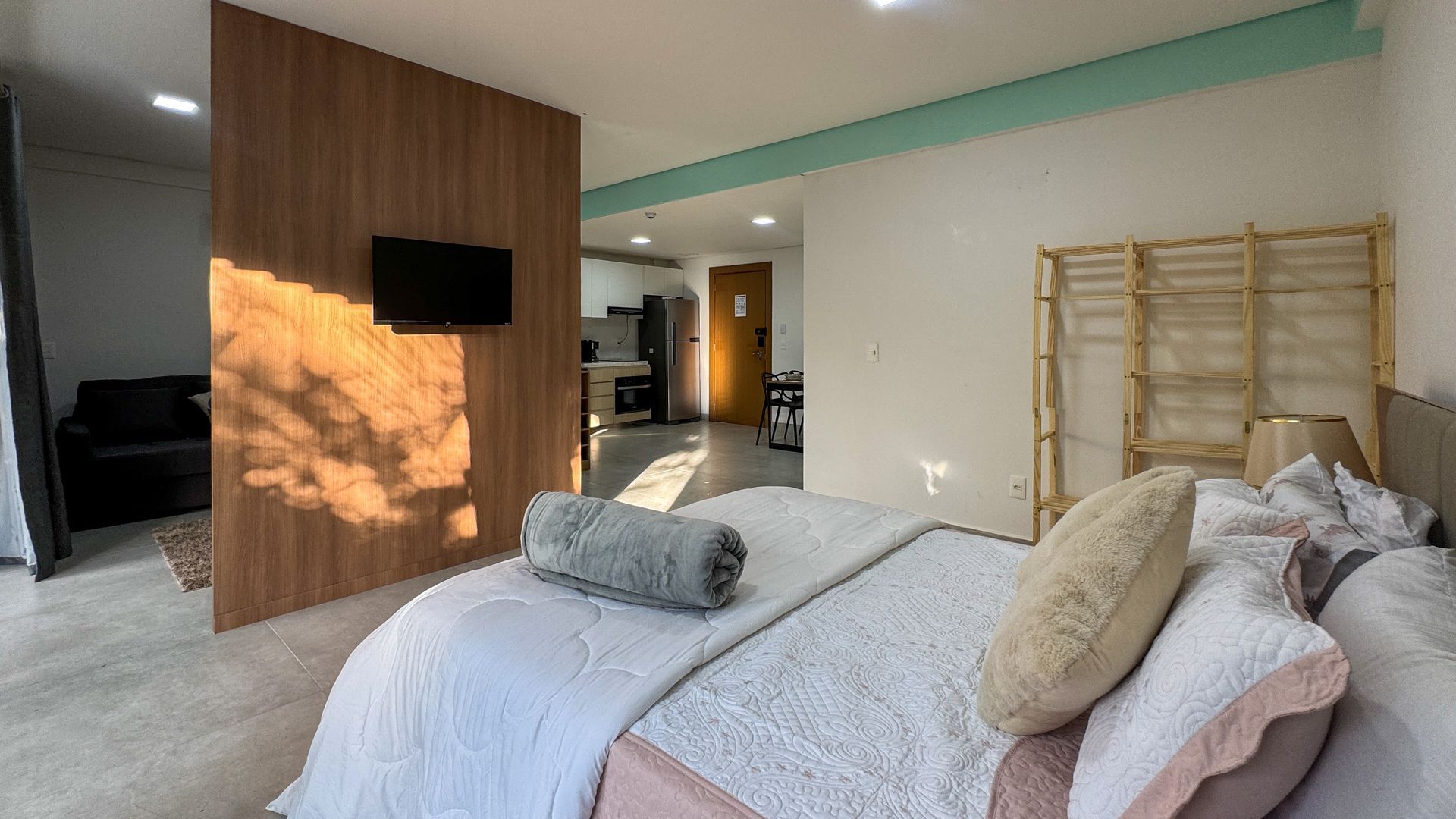 Quarto com cama, TV, painel de madeira, biombo de bambu; luz natural e decoração moderna.