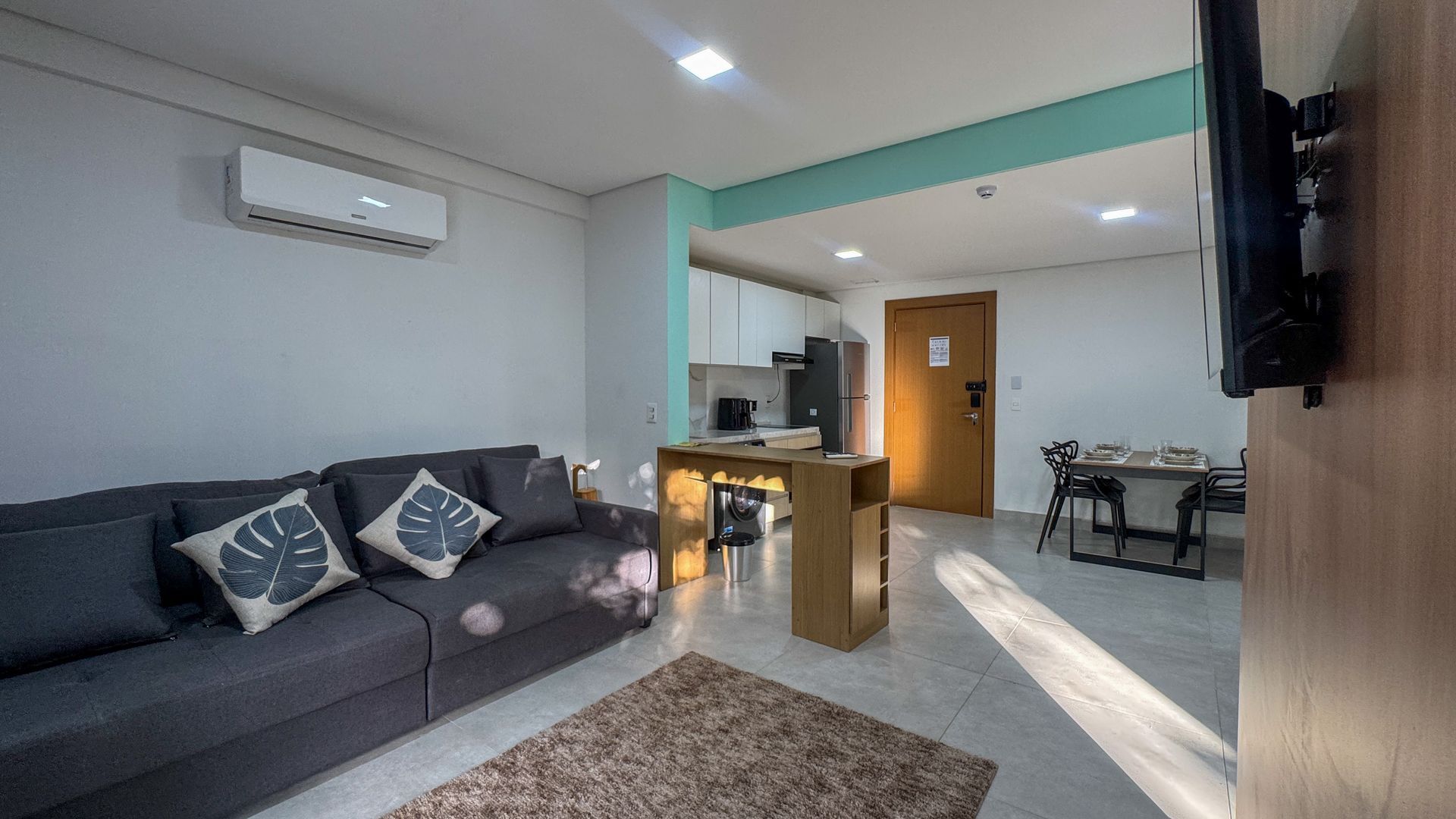 Sala de estar com sofá cinza, cozinha pequena e área de jantar.