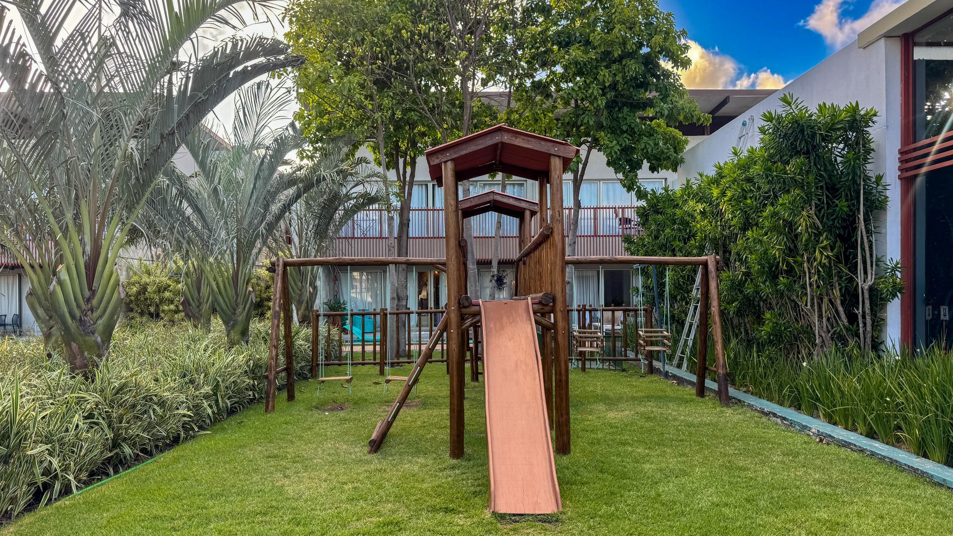 Parque infantil com estrutura de madeira, escorregador e balanços em grama verde, ao lado de um prédio e área verde.