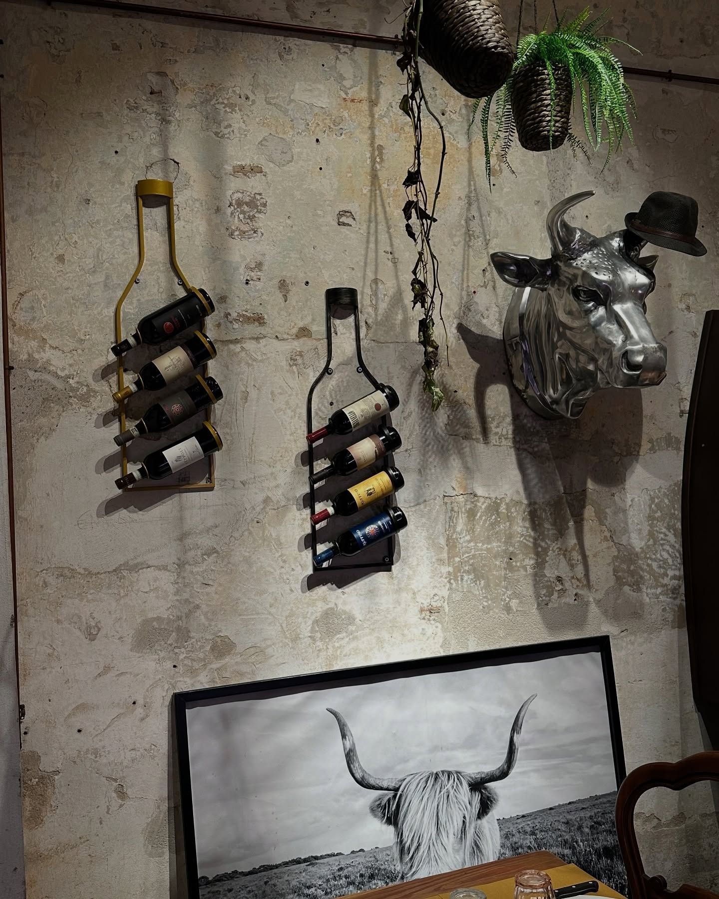 Scaffali a forma di bottiglie di vino che sostengono le bottiglie su una parete strutturata, con una scultura a forma di testa di mucca, piante e una foto incorniciata.