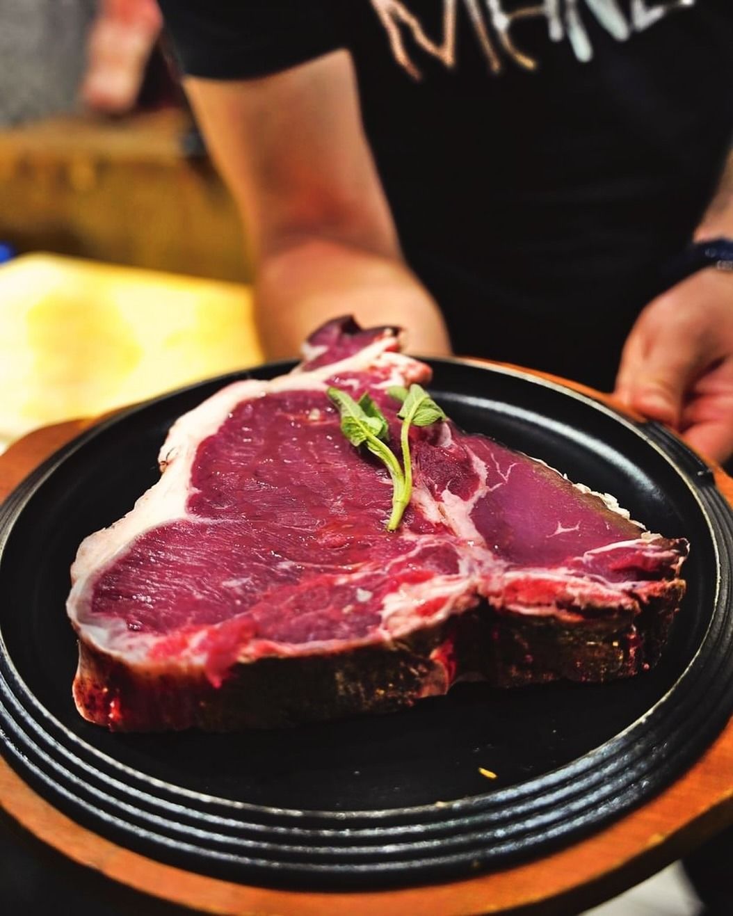 Un cameriere tiene in mano una grande bistecca di manzo cruda su un piatto nero. La carne è rossa e guarnita con erbe aromatiche verdi.
