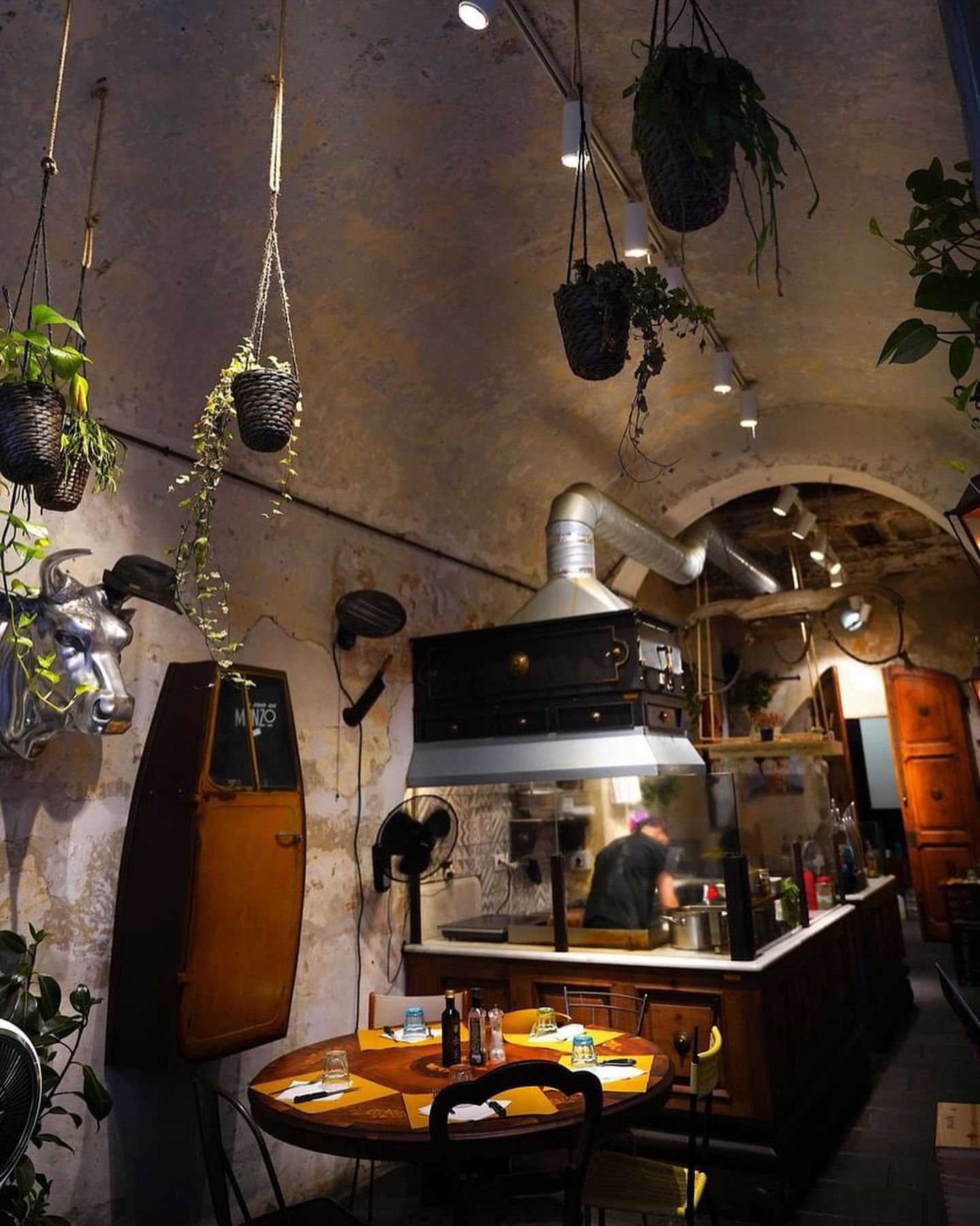 Interno di un ristorante con piante sospese, soffitto curvo e un cuoco che lavora dietro il bancone.