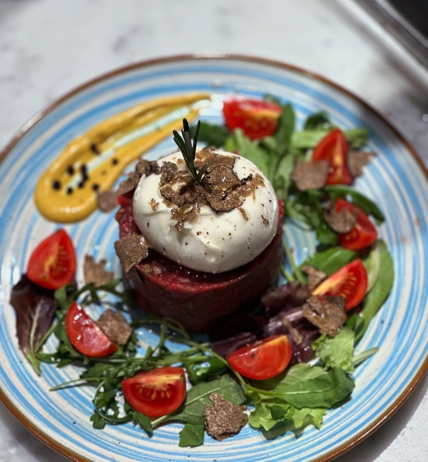 Un piatto gourmet con burrata, tartufo, pomodori e verdure su un piatto con bordo blu e salsa.