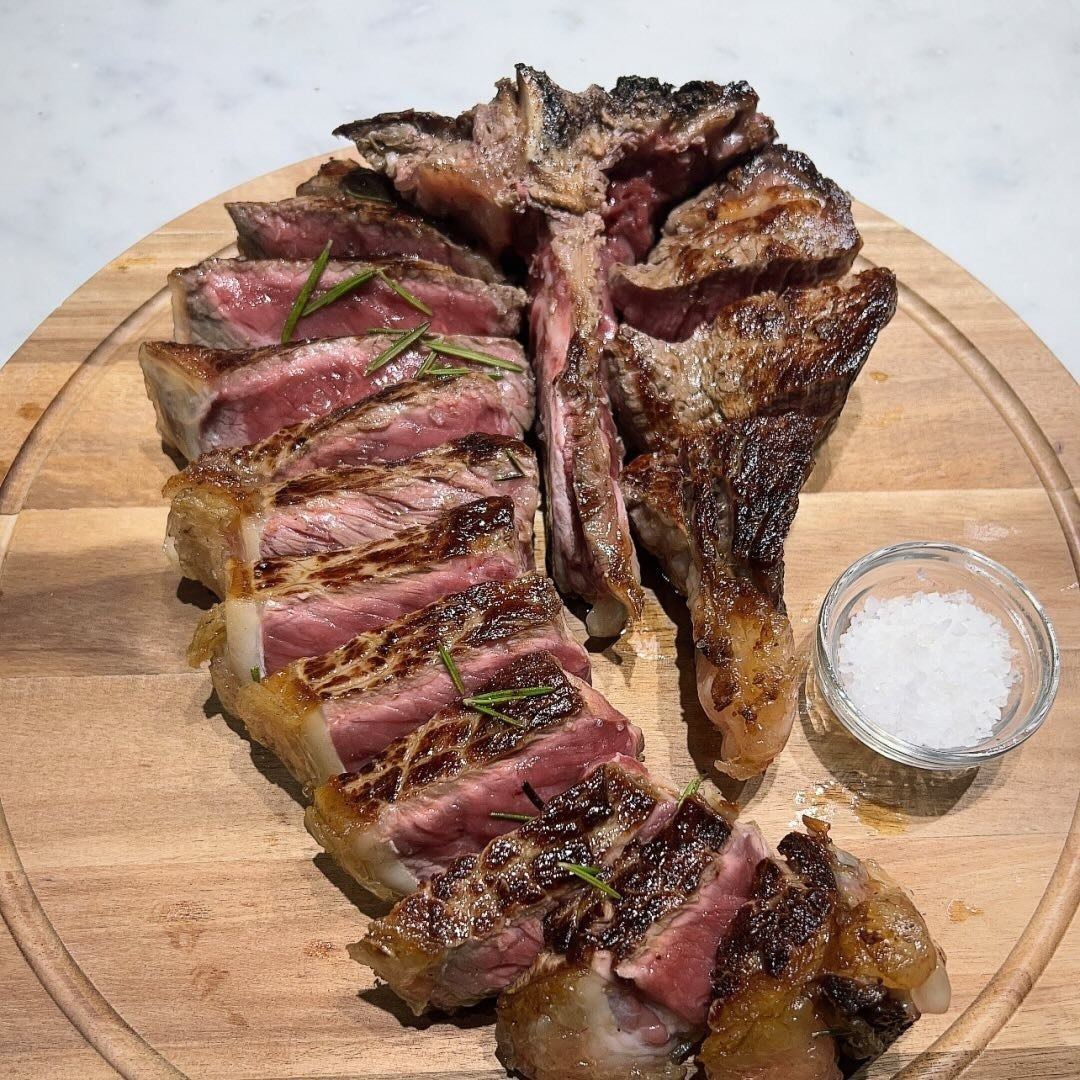 Bistecca di manzo alla fiorentina tagliata a fette e grigliata con rosmarino, servita su un tagliere di legno con una piccola ciotola di sale.