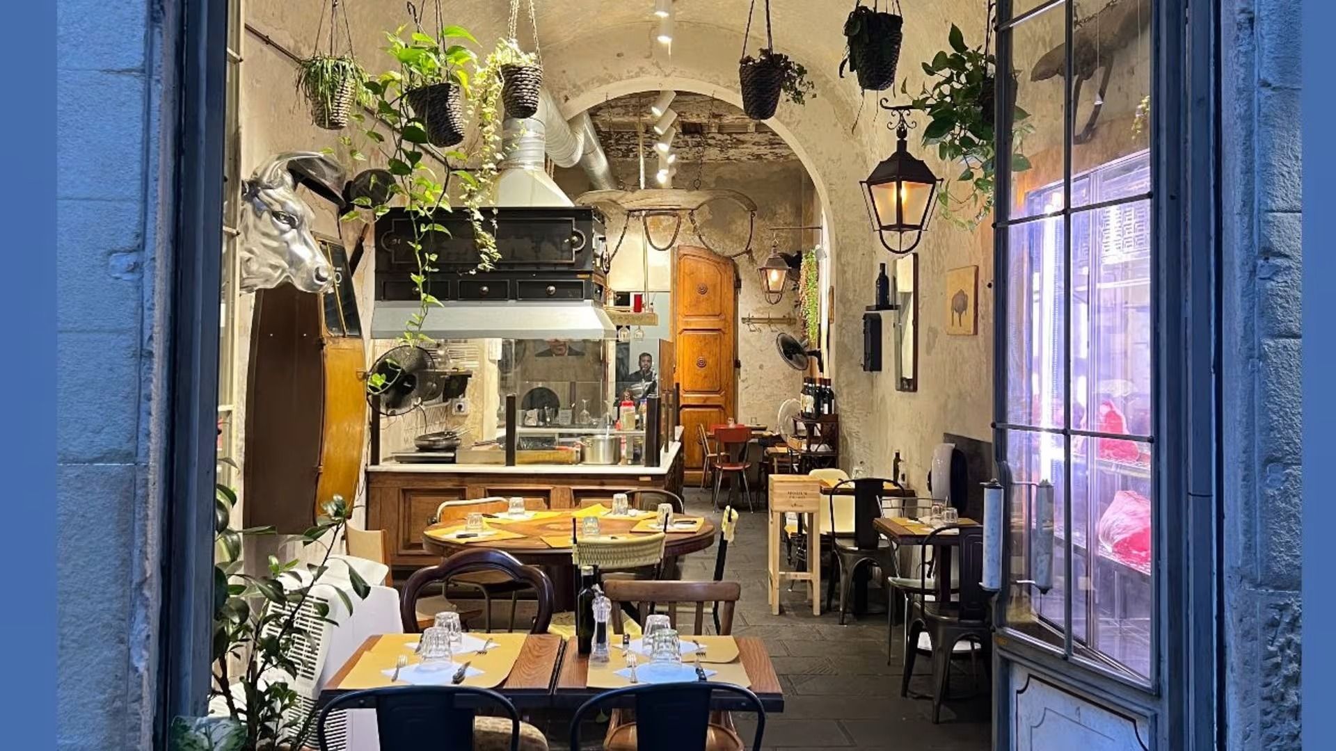 Interno del ristorante: tavoli, sedie, piante e ingresso ad arco. Luci soffuse e mattoni a vista.