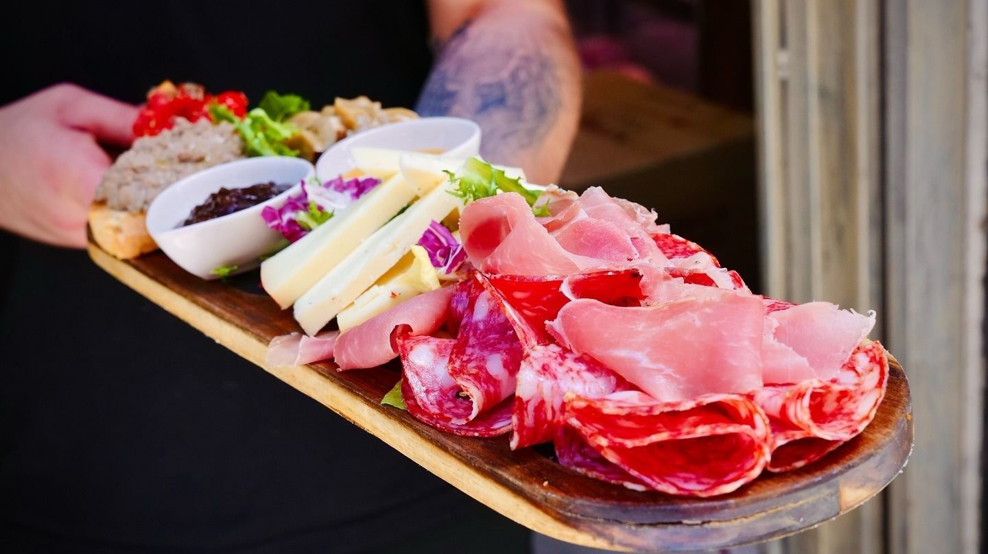 Tagliere di salumi con carni, formaggi e condimenti, tenuto in mano da una persona con un tatuaggio.