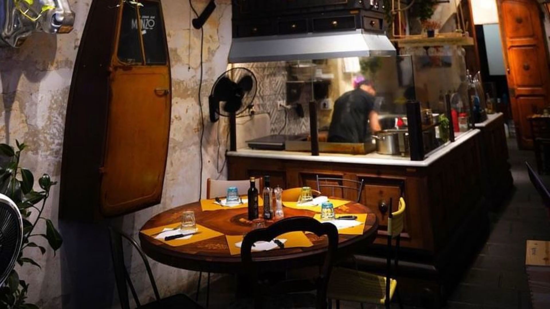 Interno del ristorante: un cuoco prepara il cibo dietro un bancone, i tavoli sono apparecchiati per mangiare.