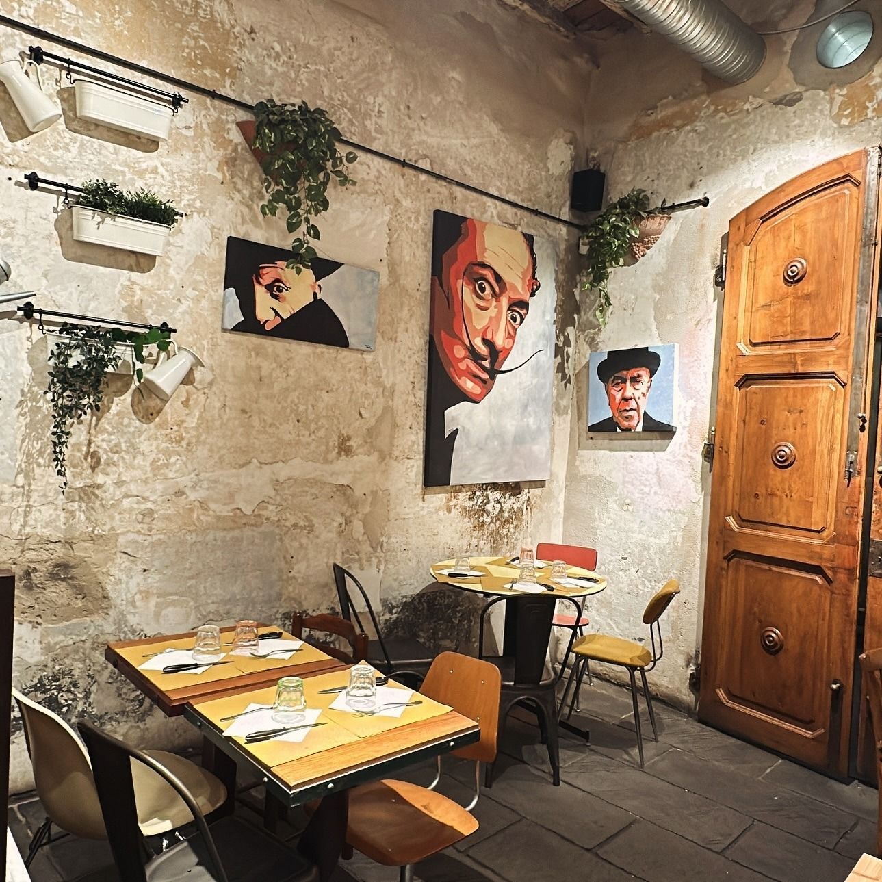 Interno di un ristorante con opere d'arte su pareti rovinate, tavoli, sedie e una porta di legno.