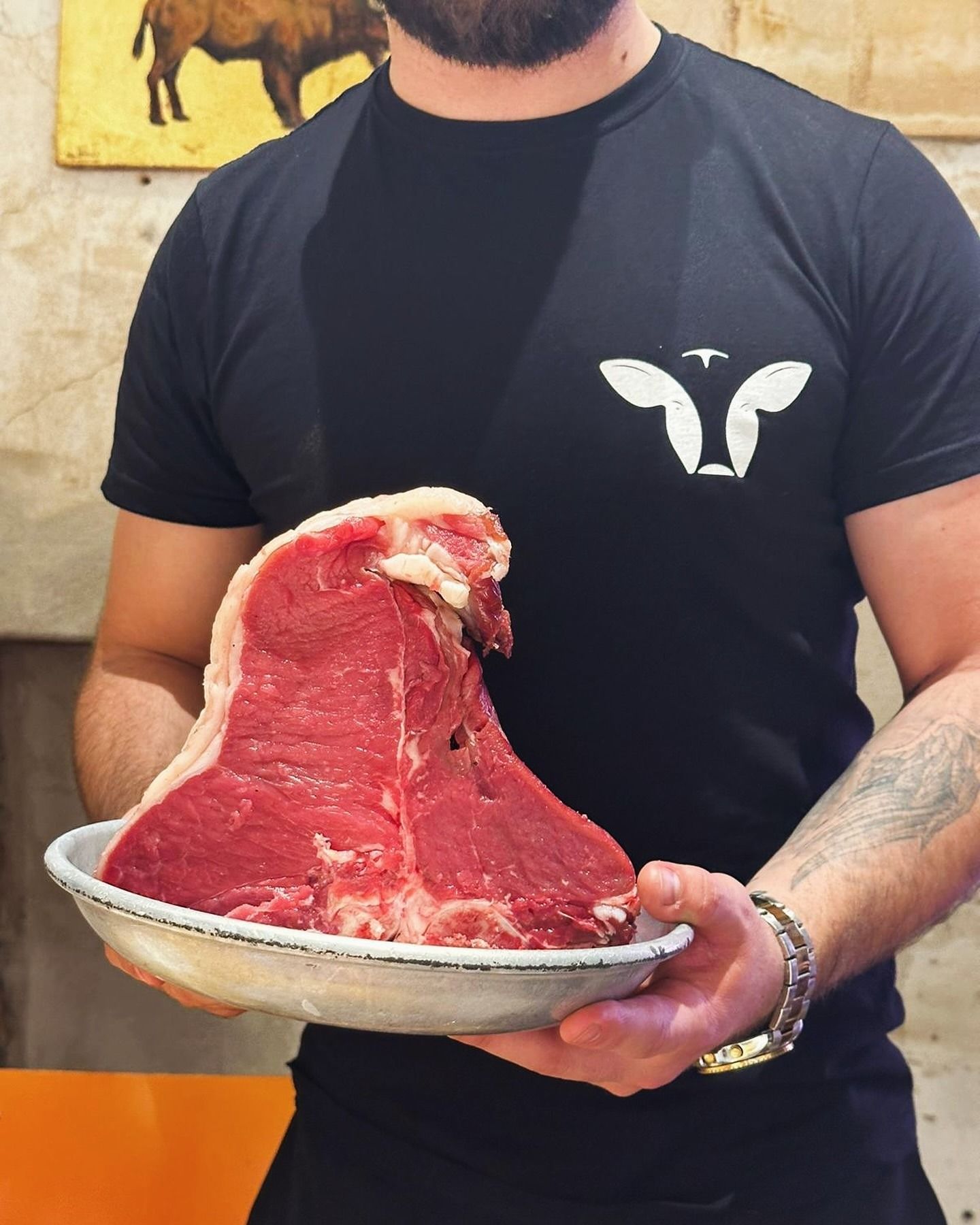 Uomo che tiene in mano una grande bistecca T-bone cruda in un piatto bianco. Indossa una maglietta nera con il logo di una mucca.