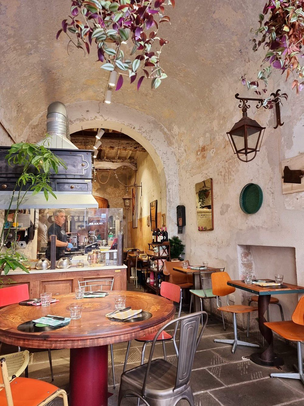 Accogliente ristorante con tavoli rotondi in legno, sedie colorate, archi e uno chef al lavoro in cucina.