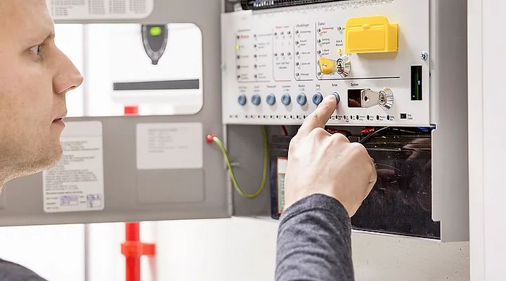 A person pressing a button on an open electronic fire alarm control panel.