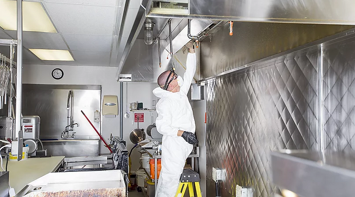 A professional in a white hazmat suit stands on a yellow stool, cleaning the interior of a stainless steel kitchen hood.