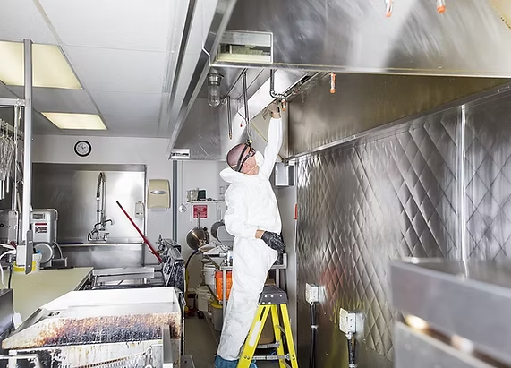 A professional in a white protective suit cleans a commercial kitchen ventilation hood while standing on a yellow ladder.
