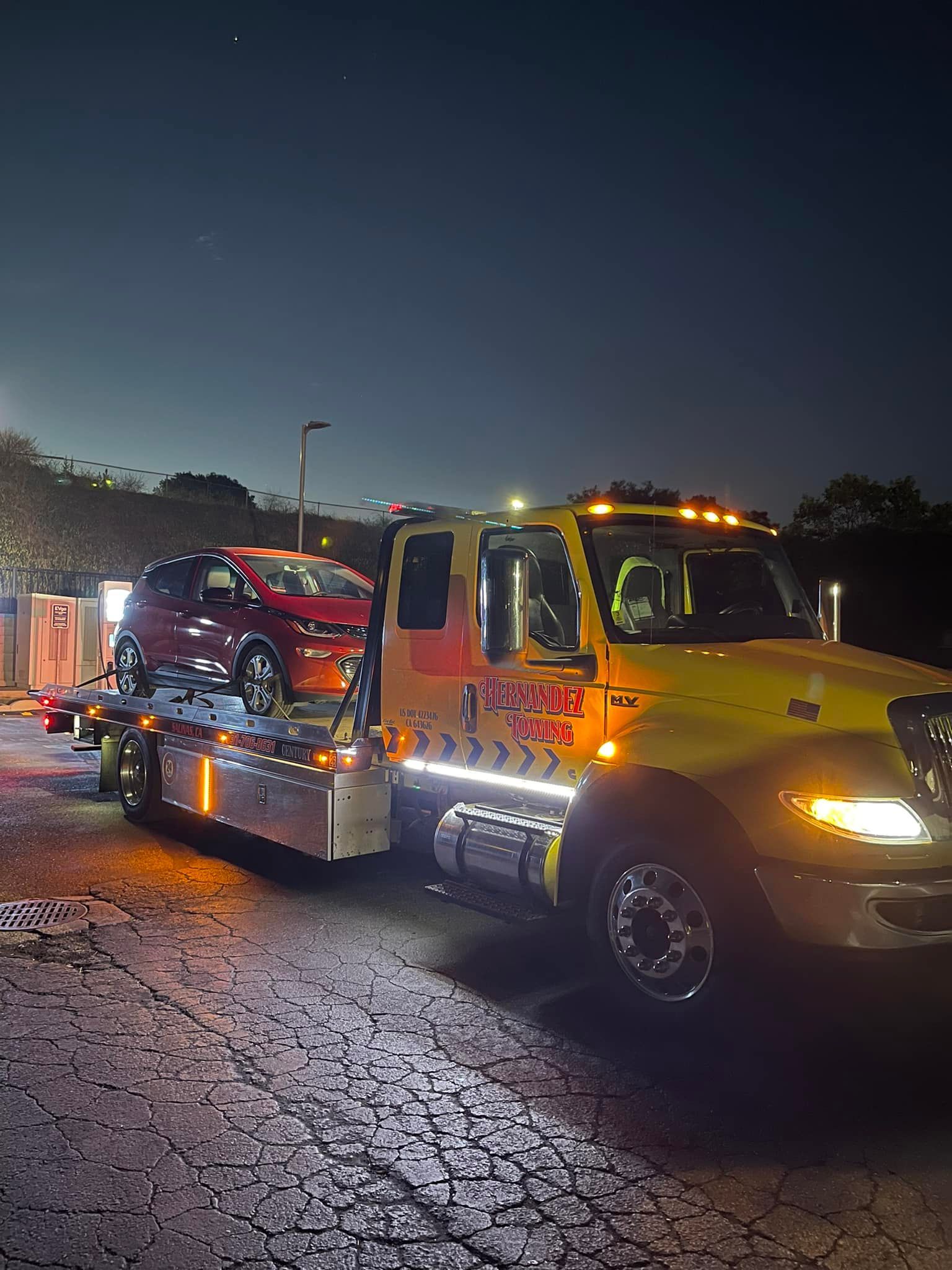 Tow truck at night transporting a red car. Yellow truck has lights on, dark sky.
