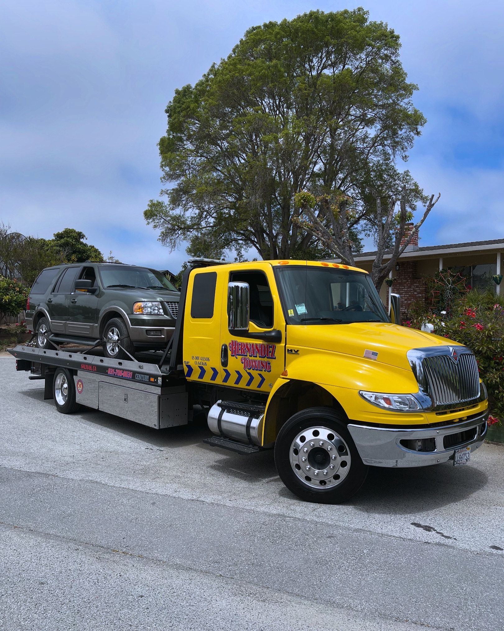 Yellow tow truck with a gray SUV on its bed, parked outside a building on a sunny day.