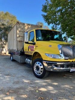 Yellow tow truck carrying a large, tan cargo container on a bright day.