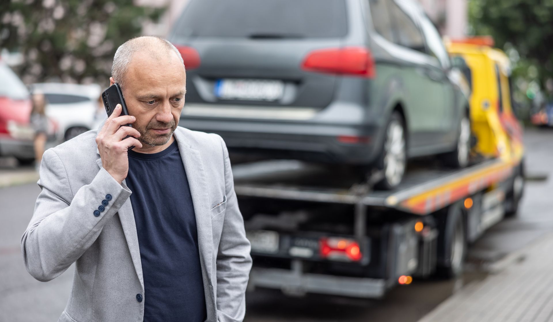 Man on phone stands near tow truck carrying his car; street setting.