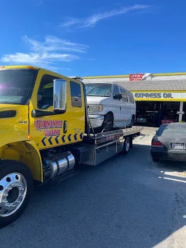 Yellow tow truck with a white van on its flatbed at an Express Oil shop on a sunny day.