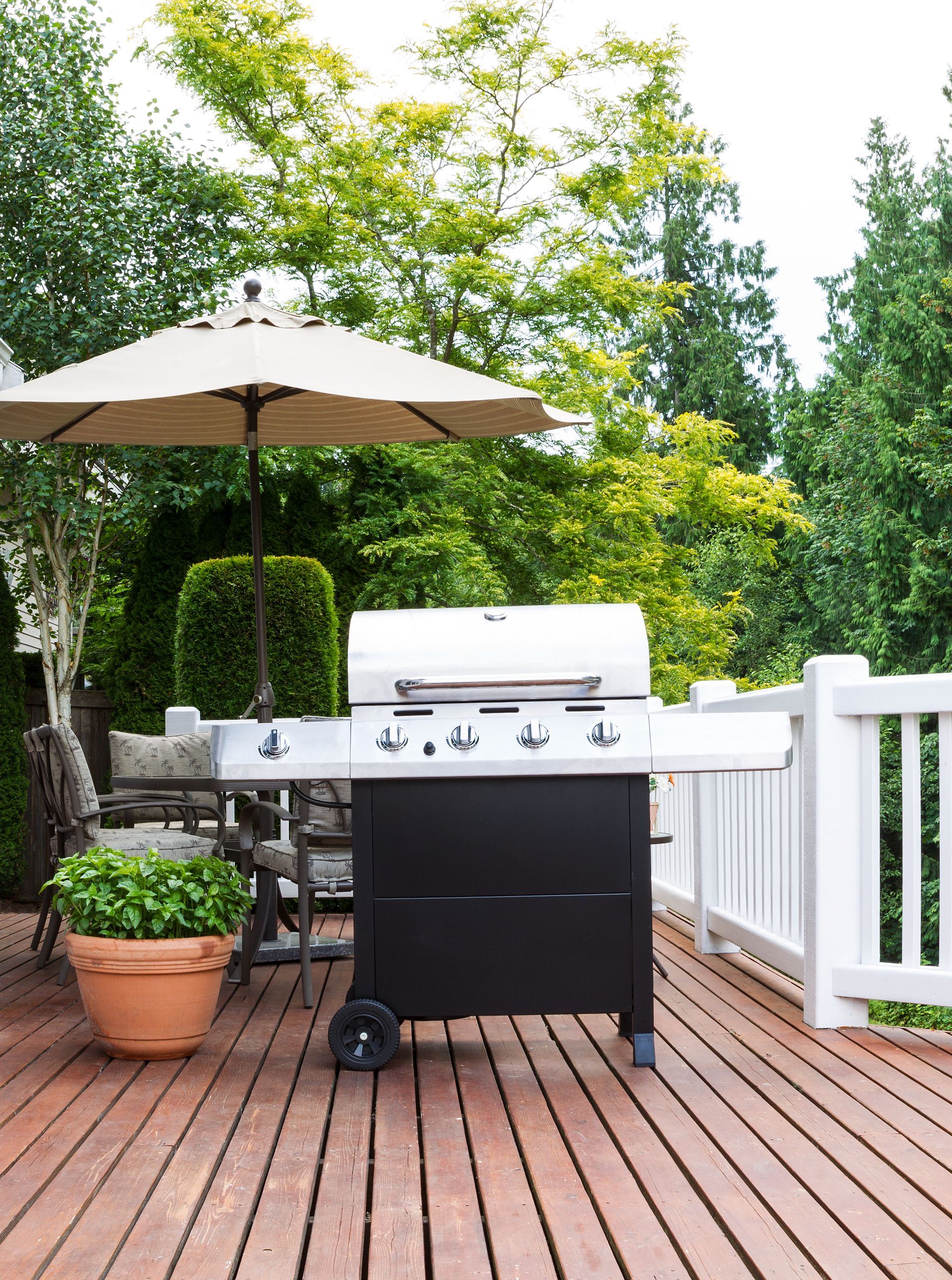 Barbecue grill on a wooden deck with an umbrella and potted plants in a backyard setting.
