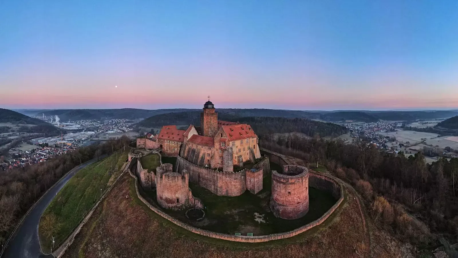 Auf einem Hügel thront eine Burg und überblickt ein Tal im Morgengrauen.
