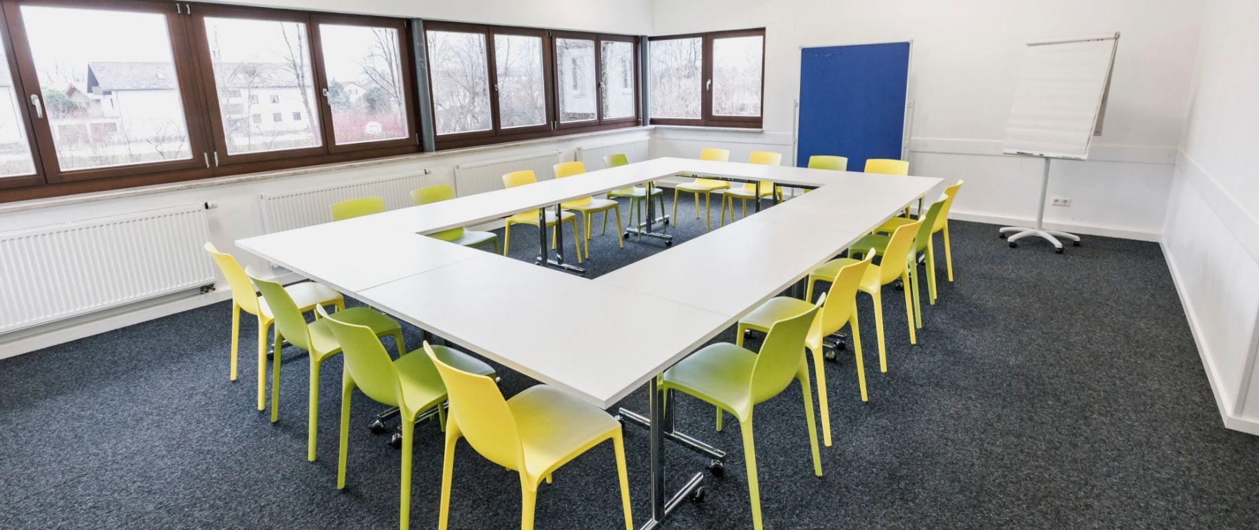 Konferenzraum mit einem rechteckigen Tisch, umgeben von gelben Stühlen, mit Fenstern und einer Whiteboard-Tafel.
