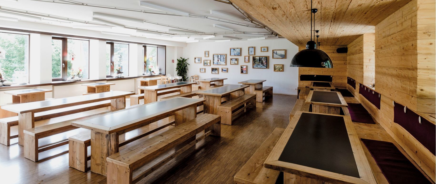 Ein hölzerner Speisesaal mit langen Tischen und Bänken, Fenstern und Bildern an den Wänden.