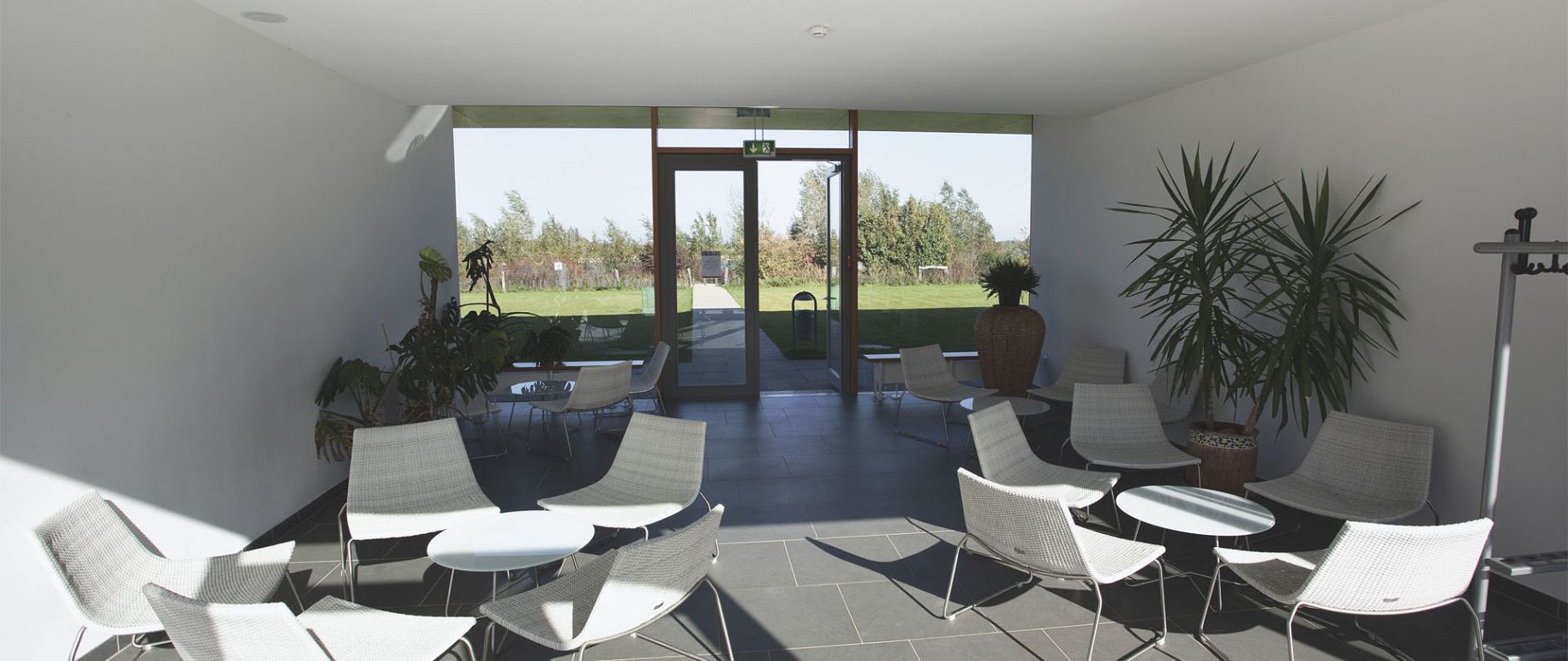 Innenbereich mit Sitzgelegenheiten, weißen Stühlen und kleinen Tischen, die auf eine offene Glastür mit Blick auf einen grünen Rasen ausgerichtet sind.