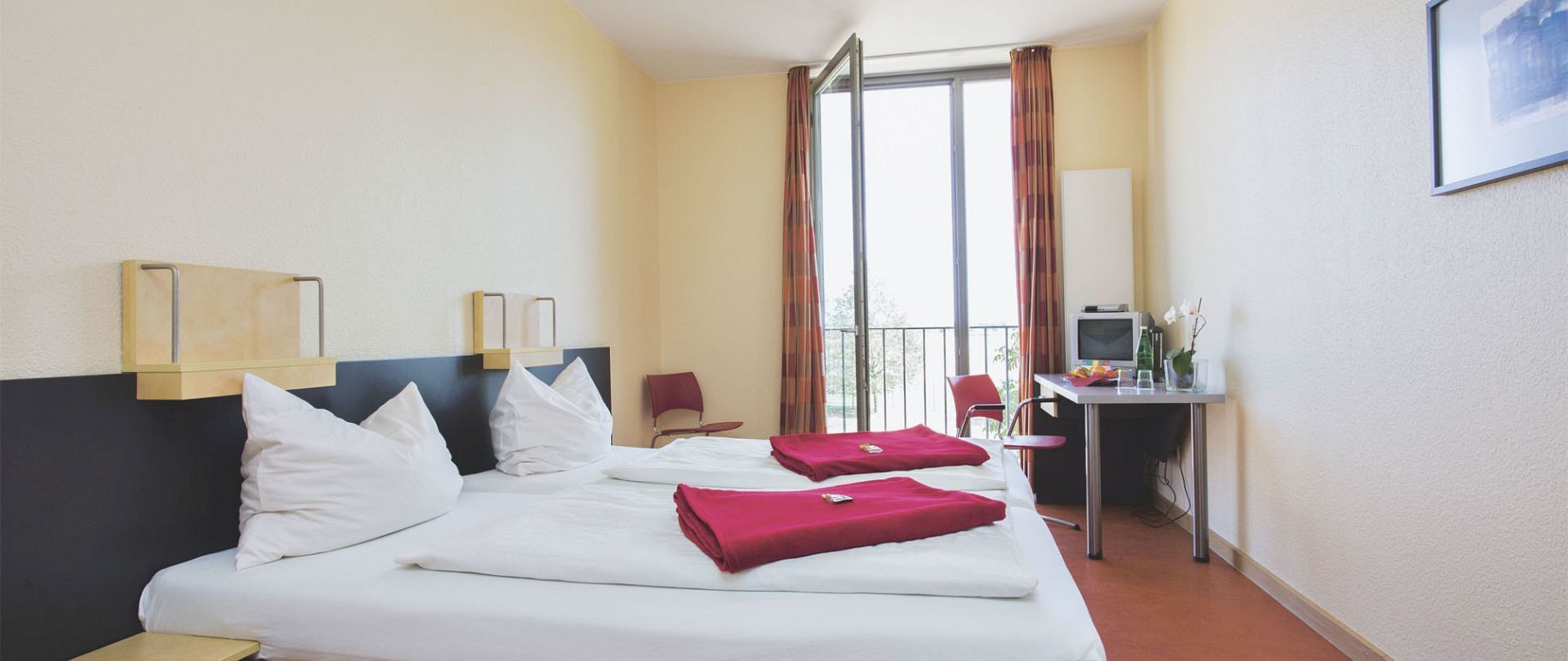 Hotelzimmer mit zwei Betten, roter Bettwäsche, Balkon und kleinem Tisch.