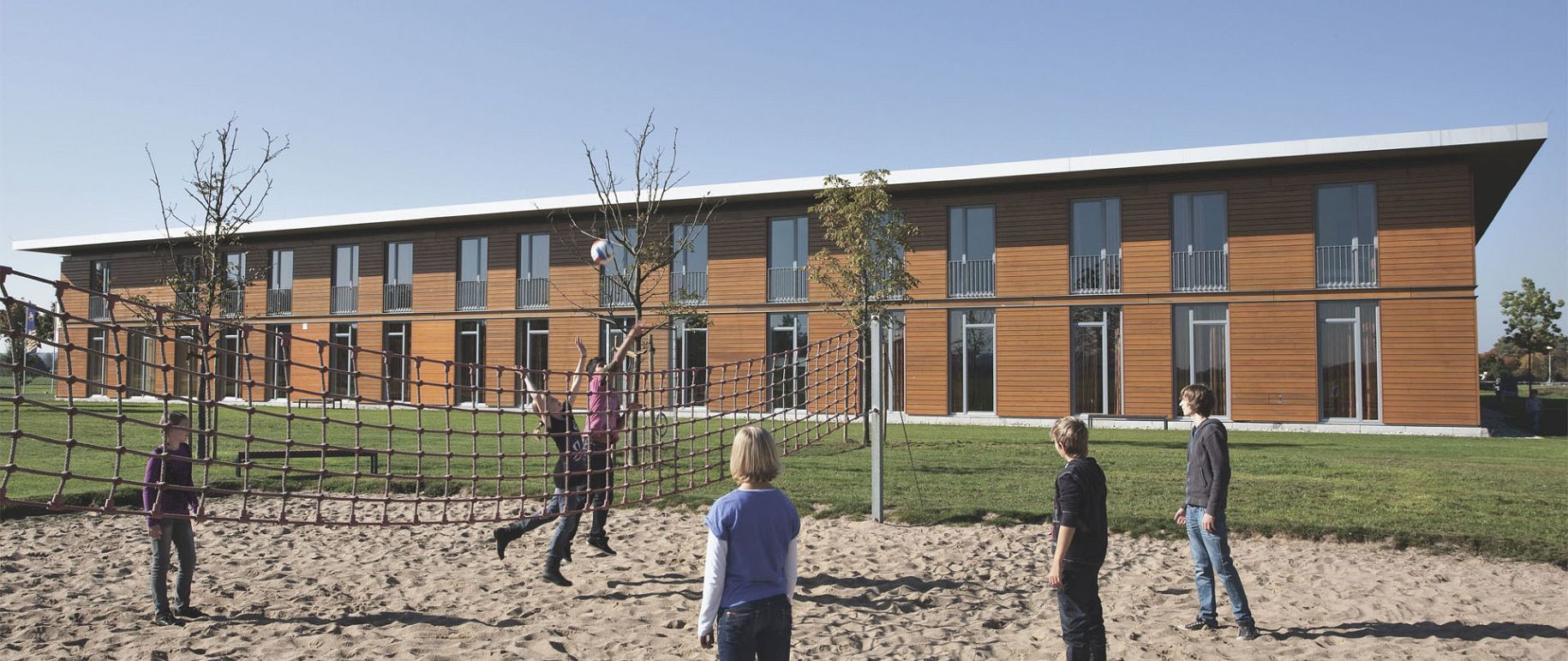 Kinder spielen auf einem Seilklettergerüst vor einem modernen, zweistöckigen Holzgebäude. Sonniger Tag.