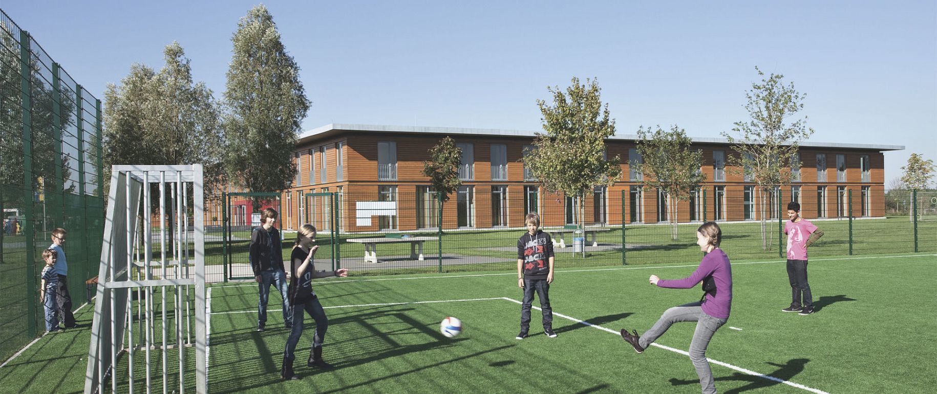 Menschen spielen Fußball auf einer grünen Wiese vor einem Gebäude. Sonniger Tag.