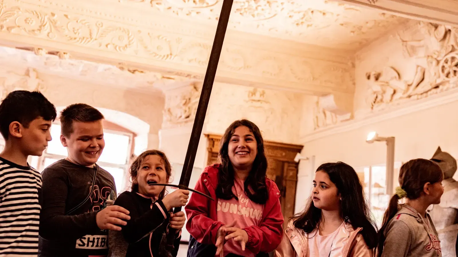 Kinder in einem Raum mit reich verzierter Decke betrachten ein langes Schwert. Ein Kind hält es in der Hand.