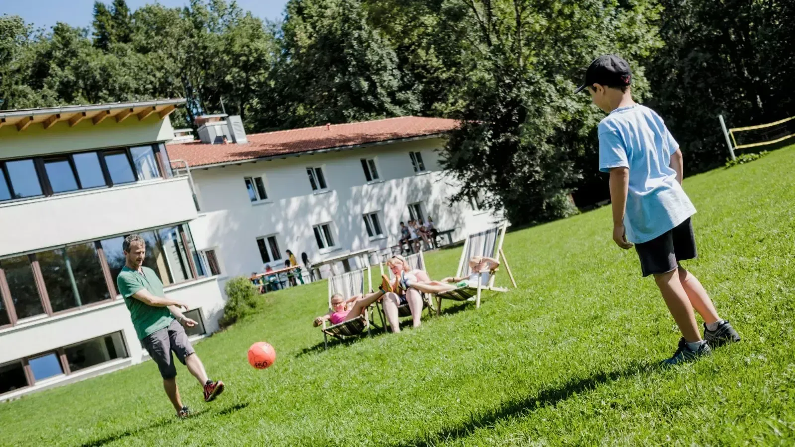 Ein Junge kickt einen Fußball auf einem grasbewachsenen Hügel; andere entspannen sich in Stühlen in der Nähe des Gebäudes.
